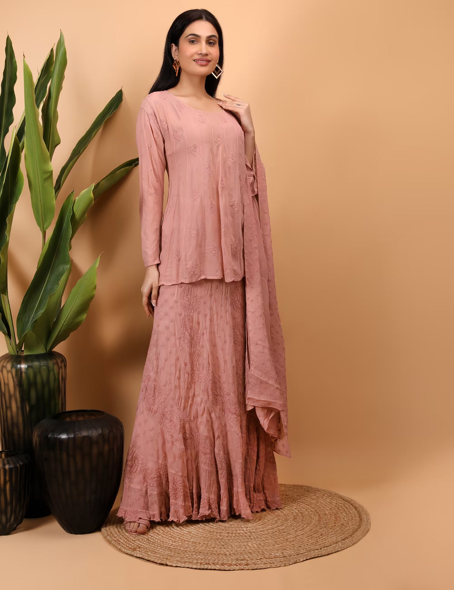 Woman in a peach traditional outfit standing against a beige background with plants.