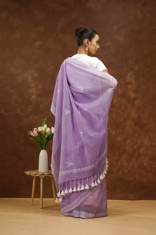 Woman wearing a purple saree with white patterns and tassels, standing against a brown background.