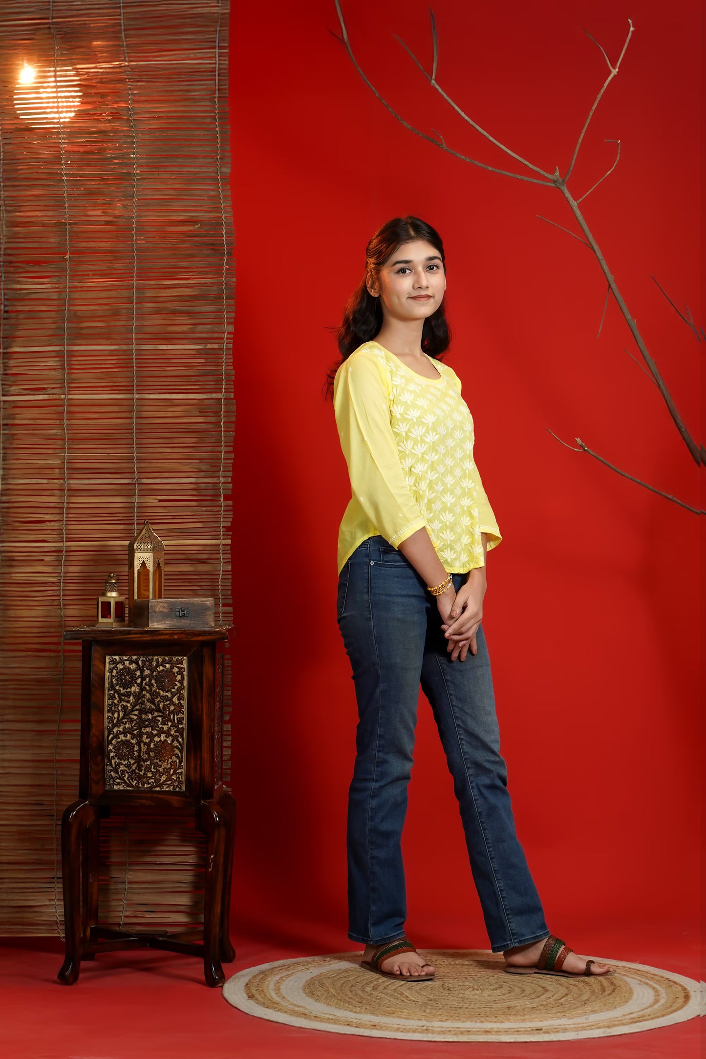 Woman in yellow top and blue jeans standing against a red wall with a decorative table and branch.