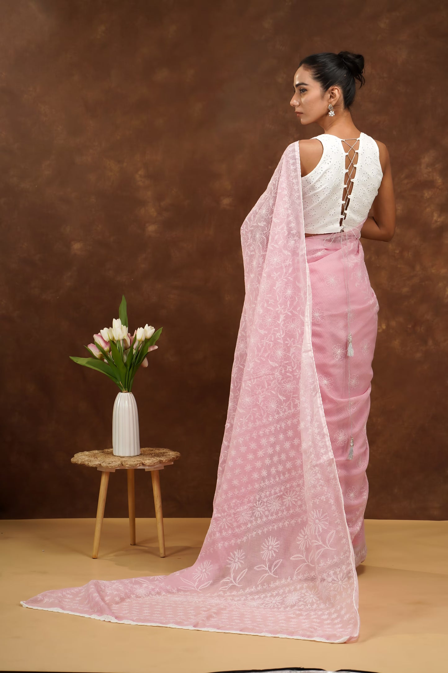 Woman in a baby pink saree with a white blouse standing against a brown background.