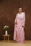 Woman in a baby pink saree standing next to a small wooden stool with a vase and flowers against a brown background