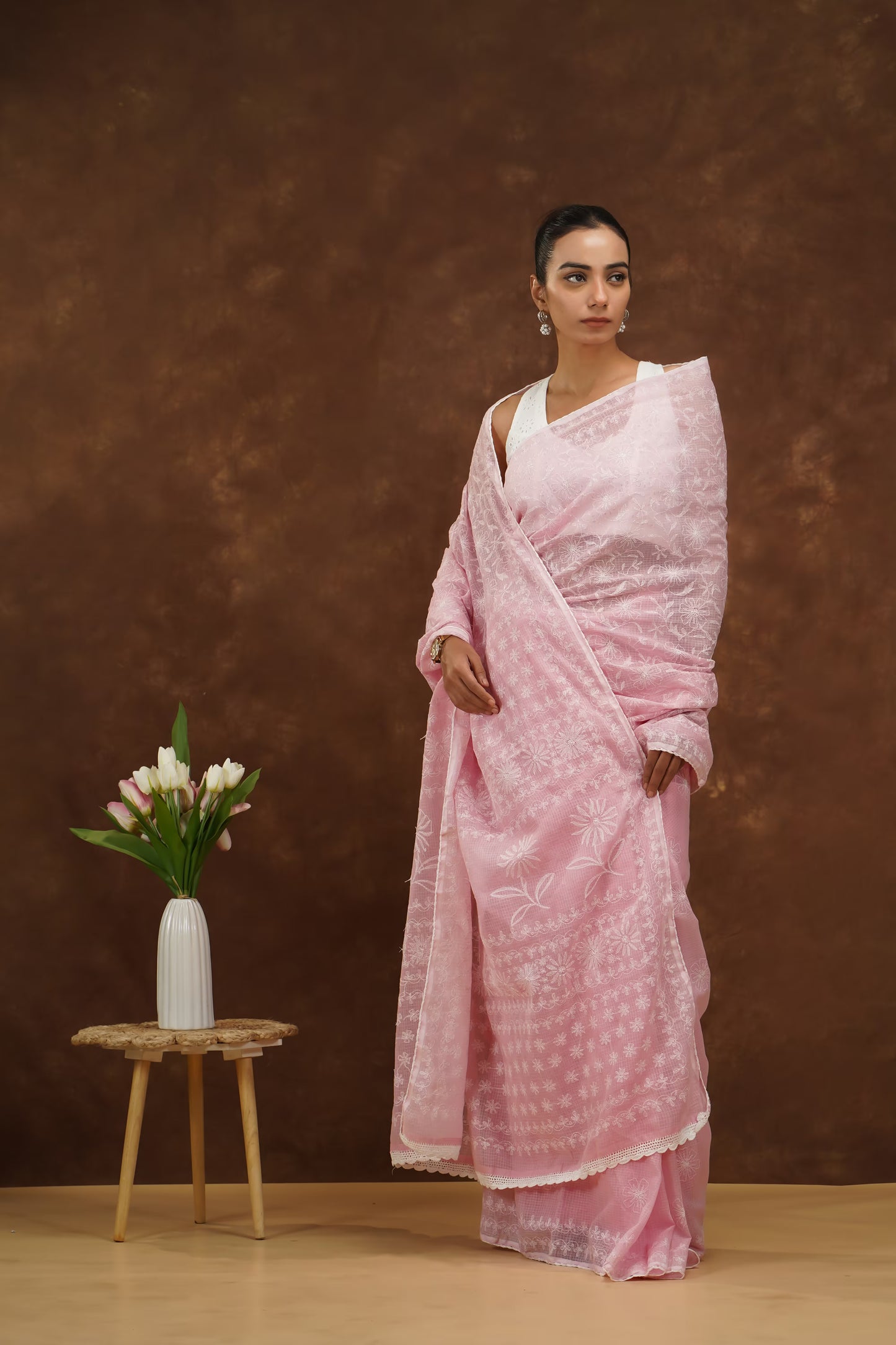Woman in a baby pink saree standing next to a small wooden stool with a vase and flowers against a brown background