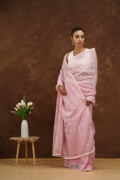 Woman in a baby pink saree standing next to a small wooden stool with a vase and flowers against a brown background