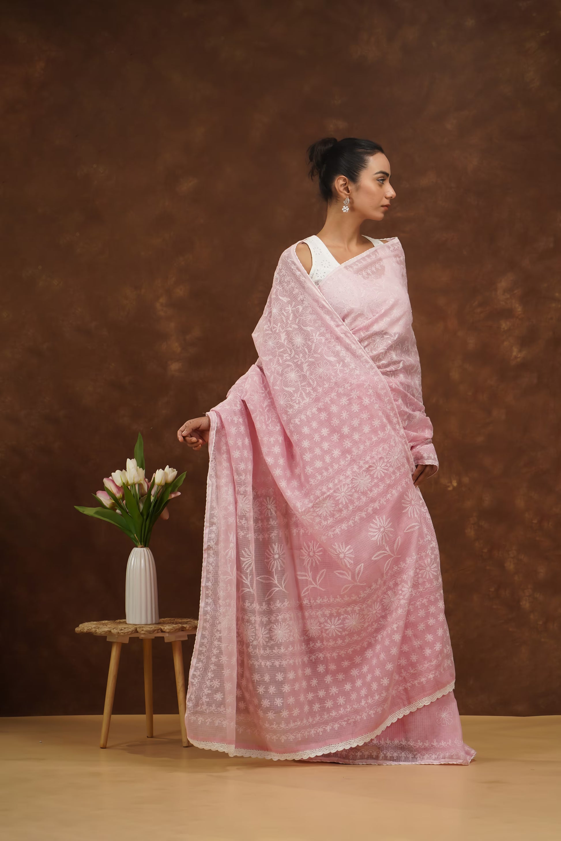 Woman in a baby pink saree standing against a brown background with a small table and flowers.