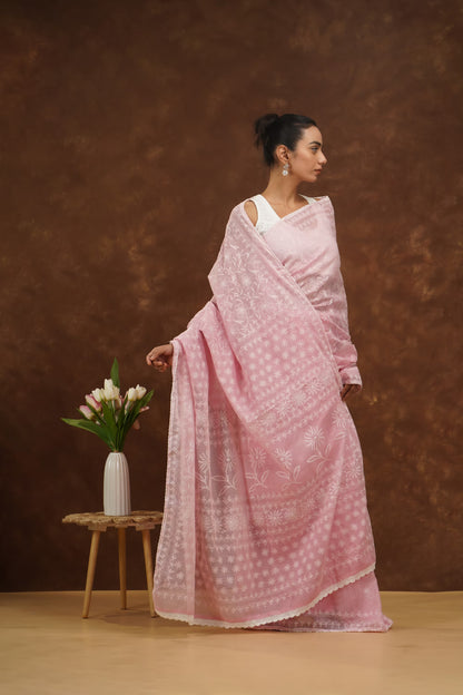 Woman in a baby pink saree standing against a brown background with a small table and flowers.