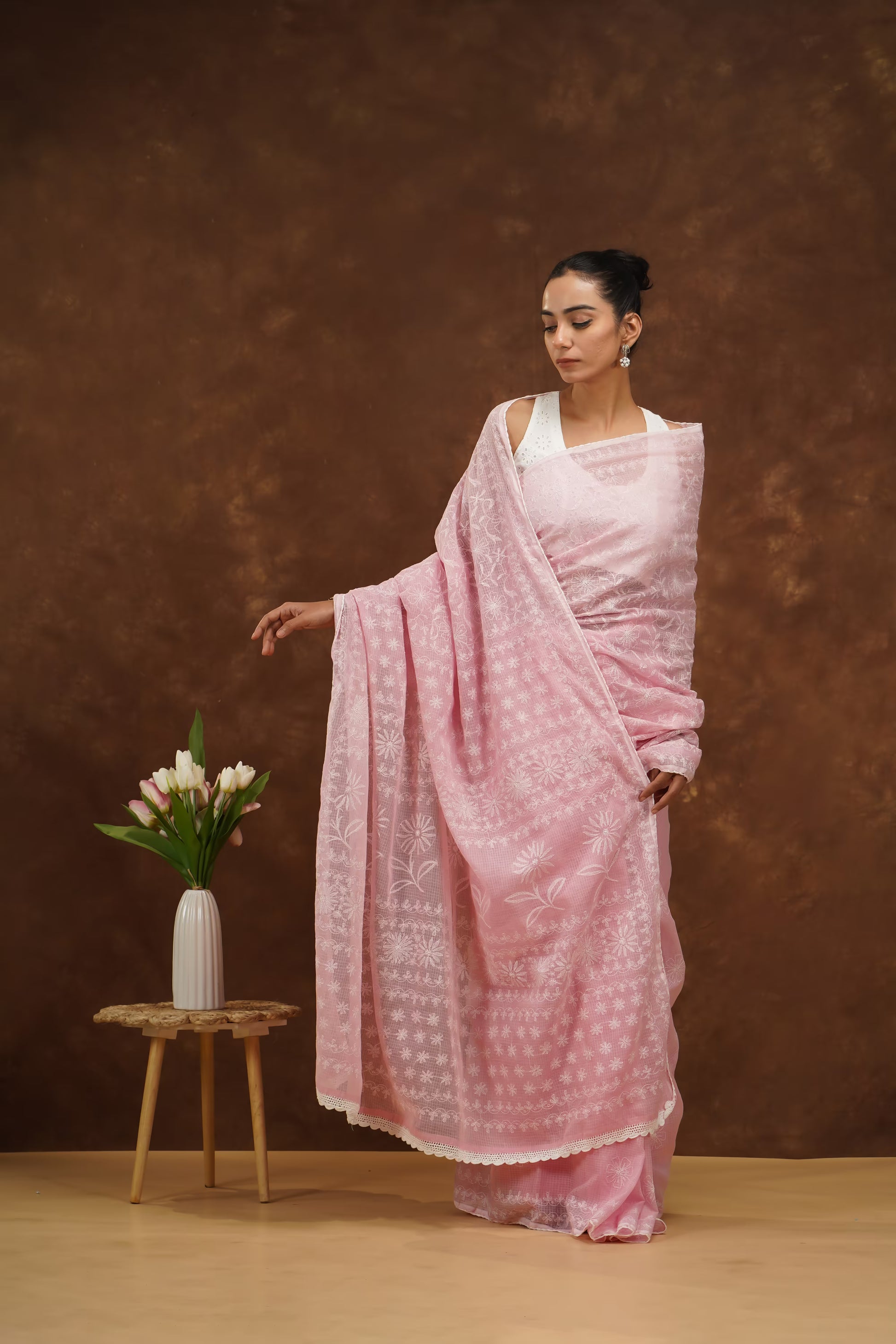 Woman in a pink saree standing next to a small table with a vase of flowers against a brown background