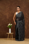 Woman in a black saree standing next to a small table with a vase of flowers against a brown background