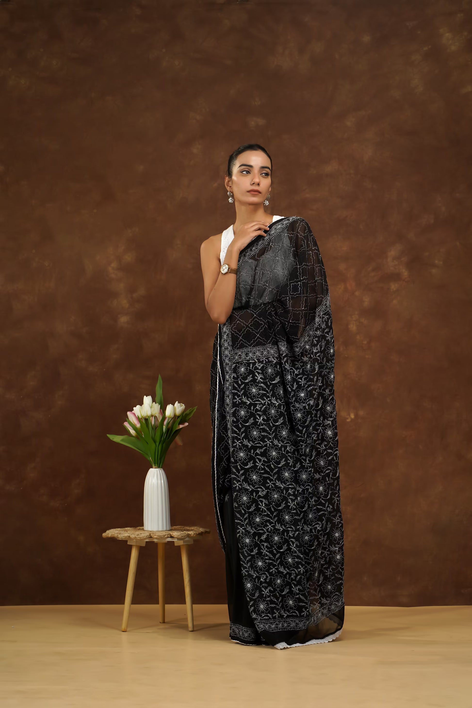 Woman in a black saree standing next to a small wooden table with a vase of flowers against a brown background
