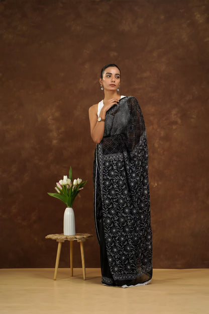Woman in a black saree standing next to a small wooden table with a vase of flowers against a brown background