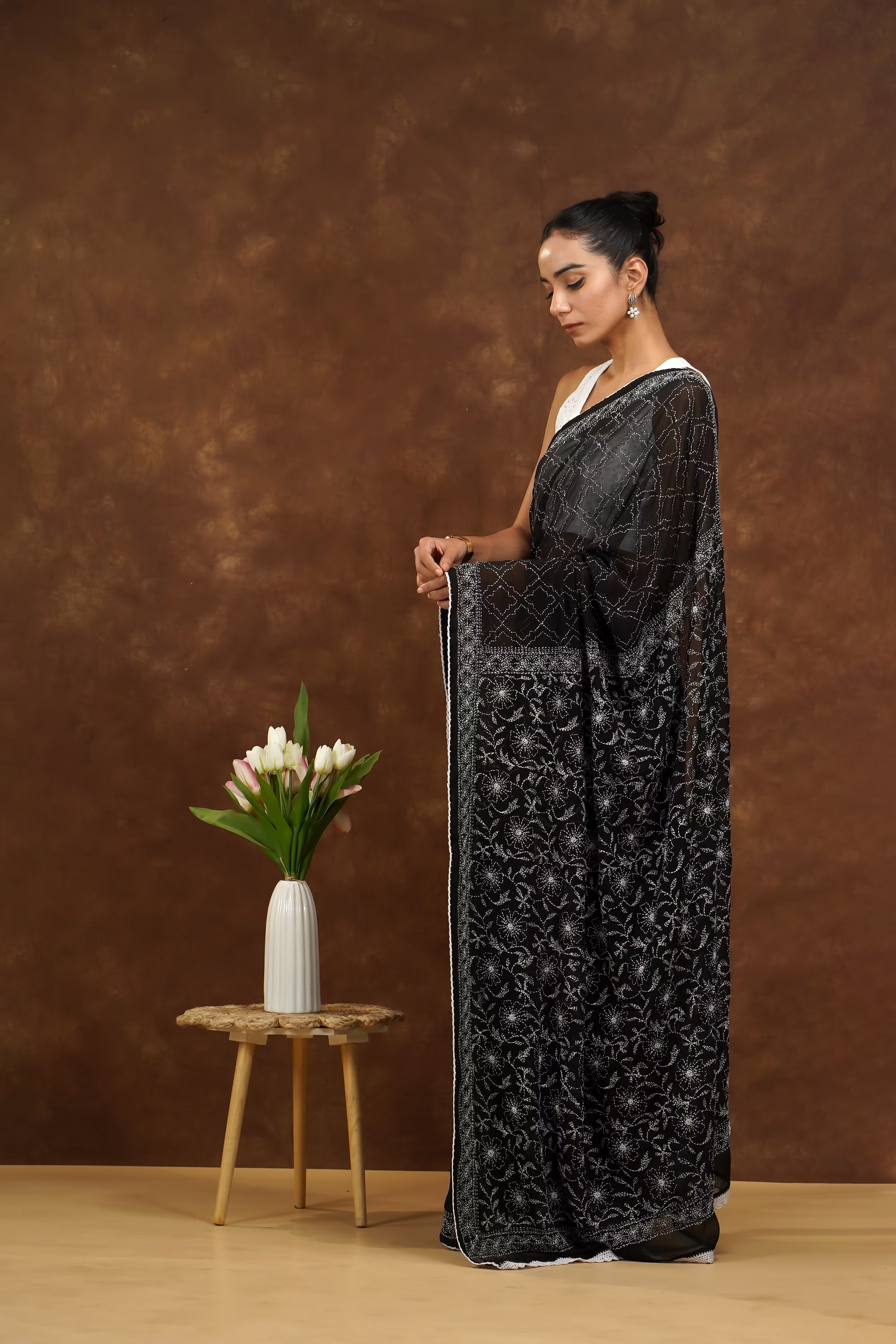 Woman in a black embroidered dress standing next to a small table with flowers against a brown background