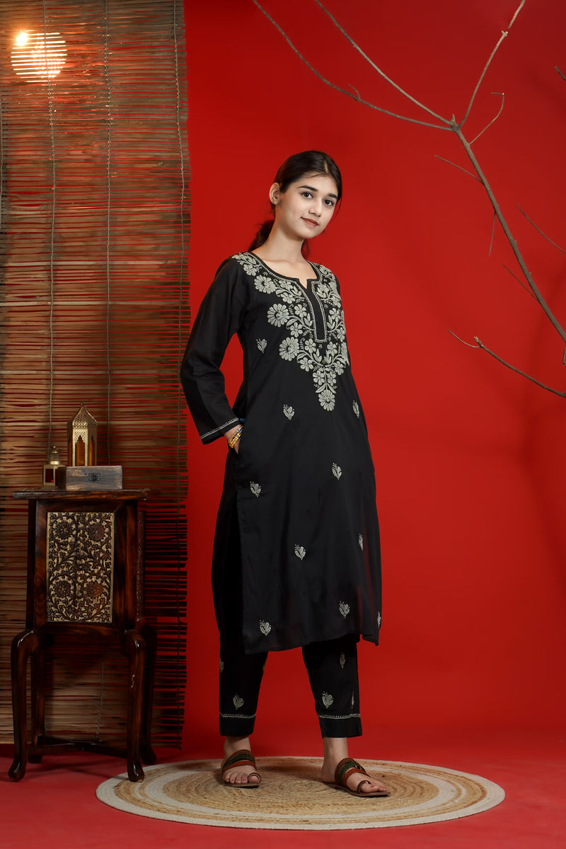 Woman in black embroidered traditional outfit standing against a red wall with a wooden side table.