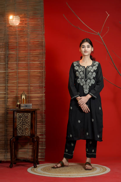 Woman in black embroidered outfit standing against a red wall with a wooden side table and decorative items.