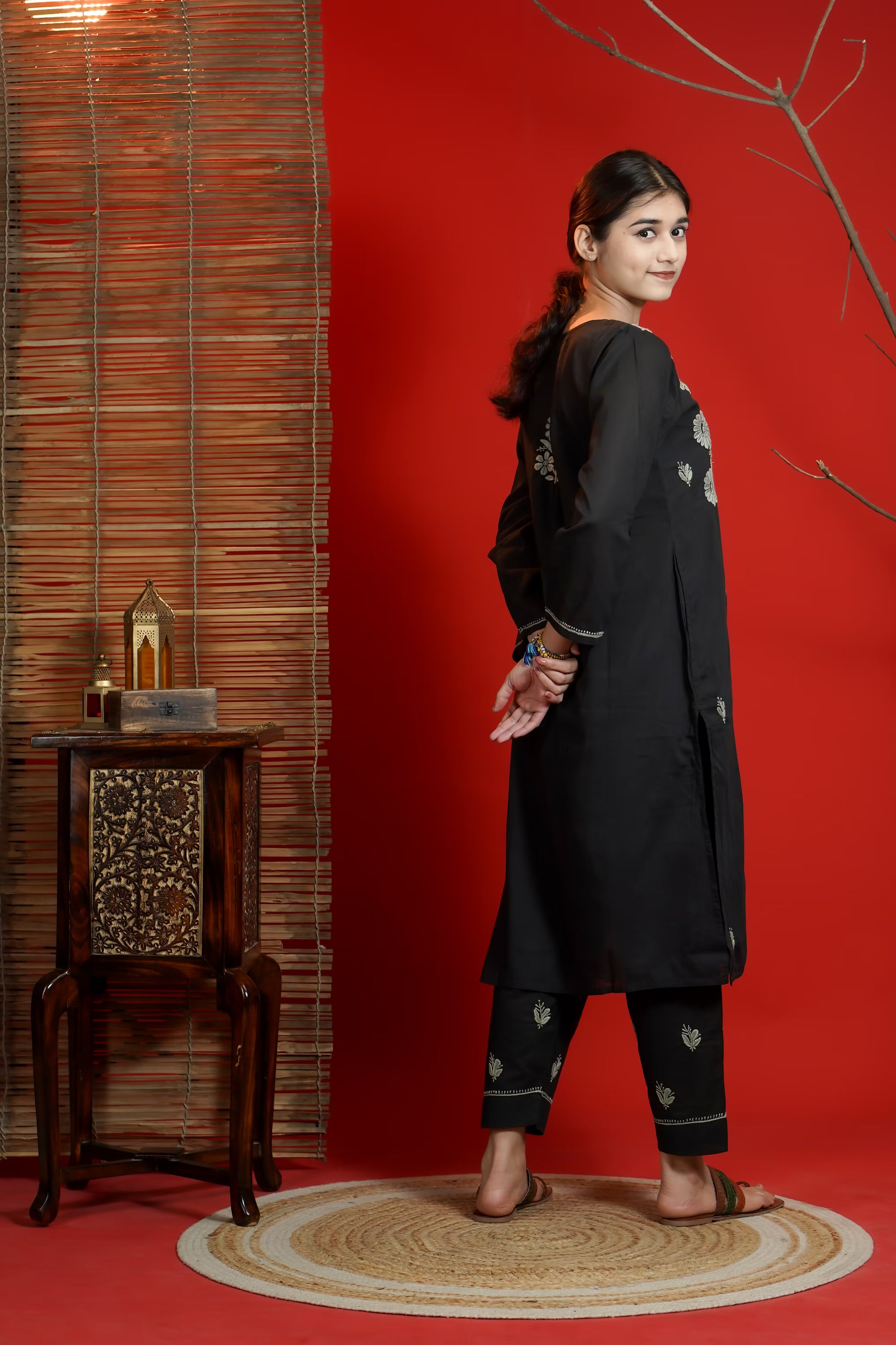 Woman in a black traditional outfit standing against a red wall with a wooden side table.