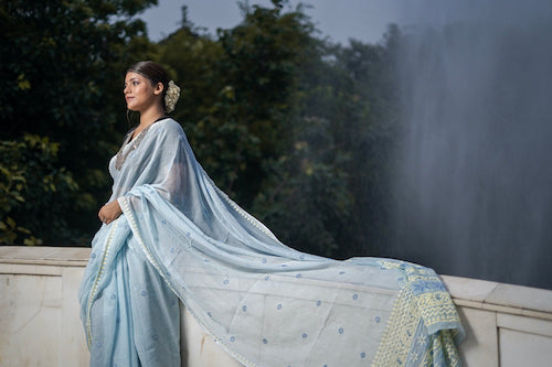 Woman in a light blue saree standing by a fountain with trees in the background