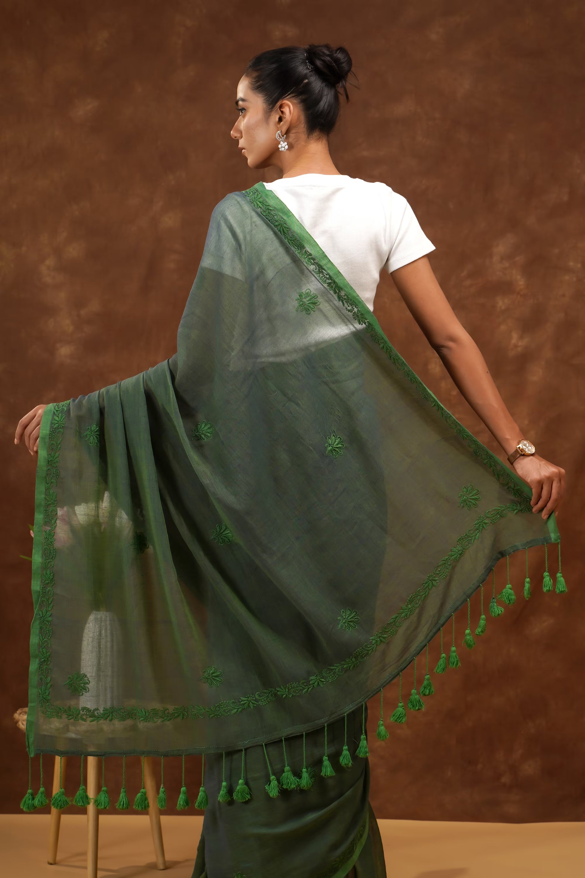 Woman wearing a dark green saree with floral patterns and tassels against a brown background