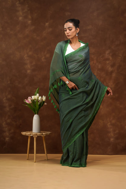 Woman in a dark green saree standing next to a small table with flowers against a brown background