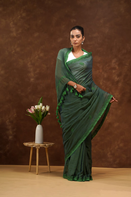 Woman in a dark green saree standing next to a small table with flowers against a brown background