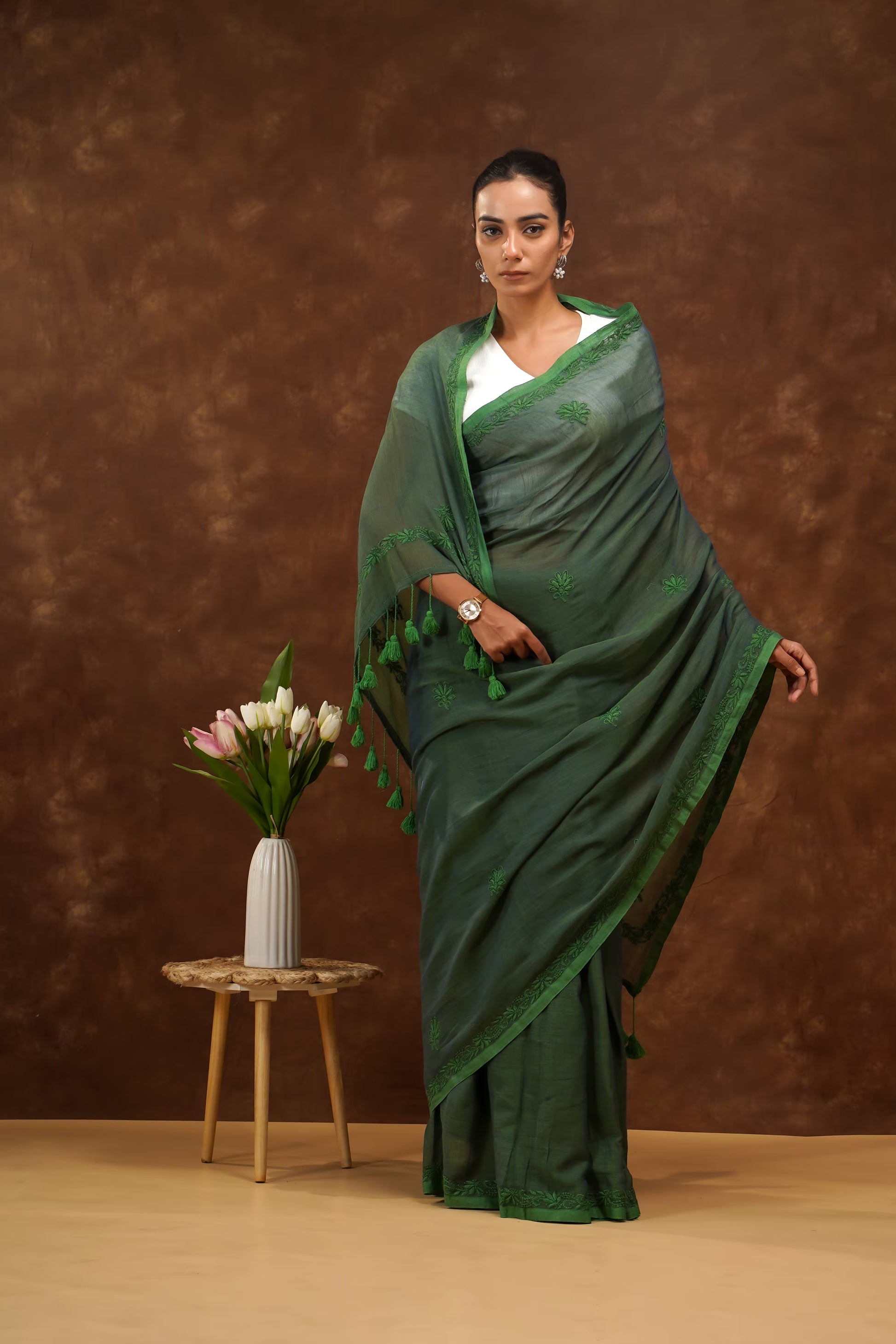 Woman in a dark green saree standing next to a small table with a vase of flowers against a brown background 