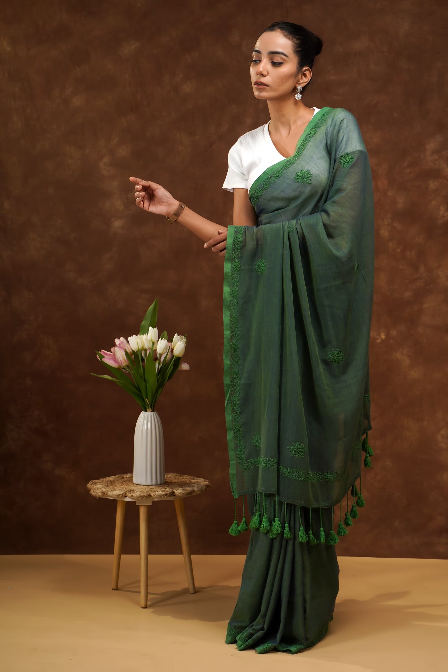 Woman in a dark green saree with white blouse standing next to a small table with flowers against a brown background