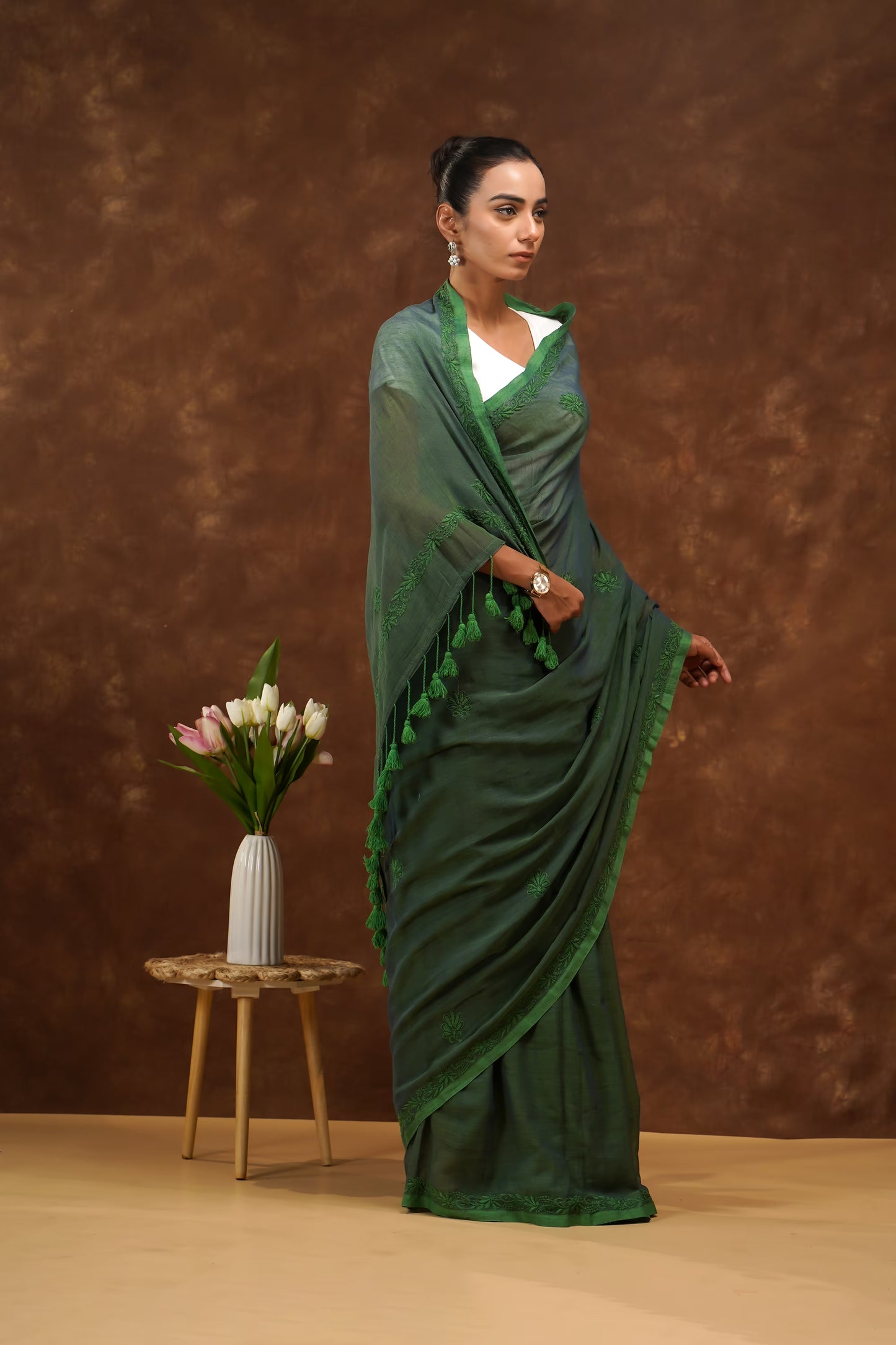 Woman in a dark green saree standing against a brown background with a small table and flowers.