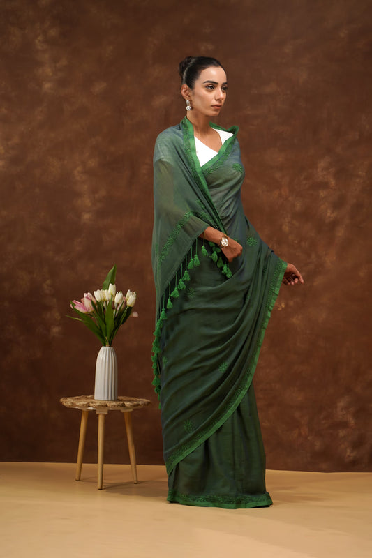 Woman in a dark green saree standing against a brown background with a small table and flowers.