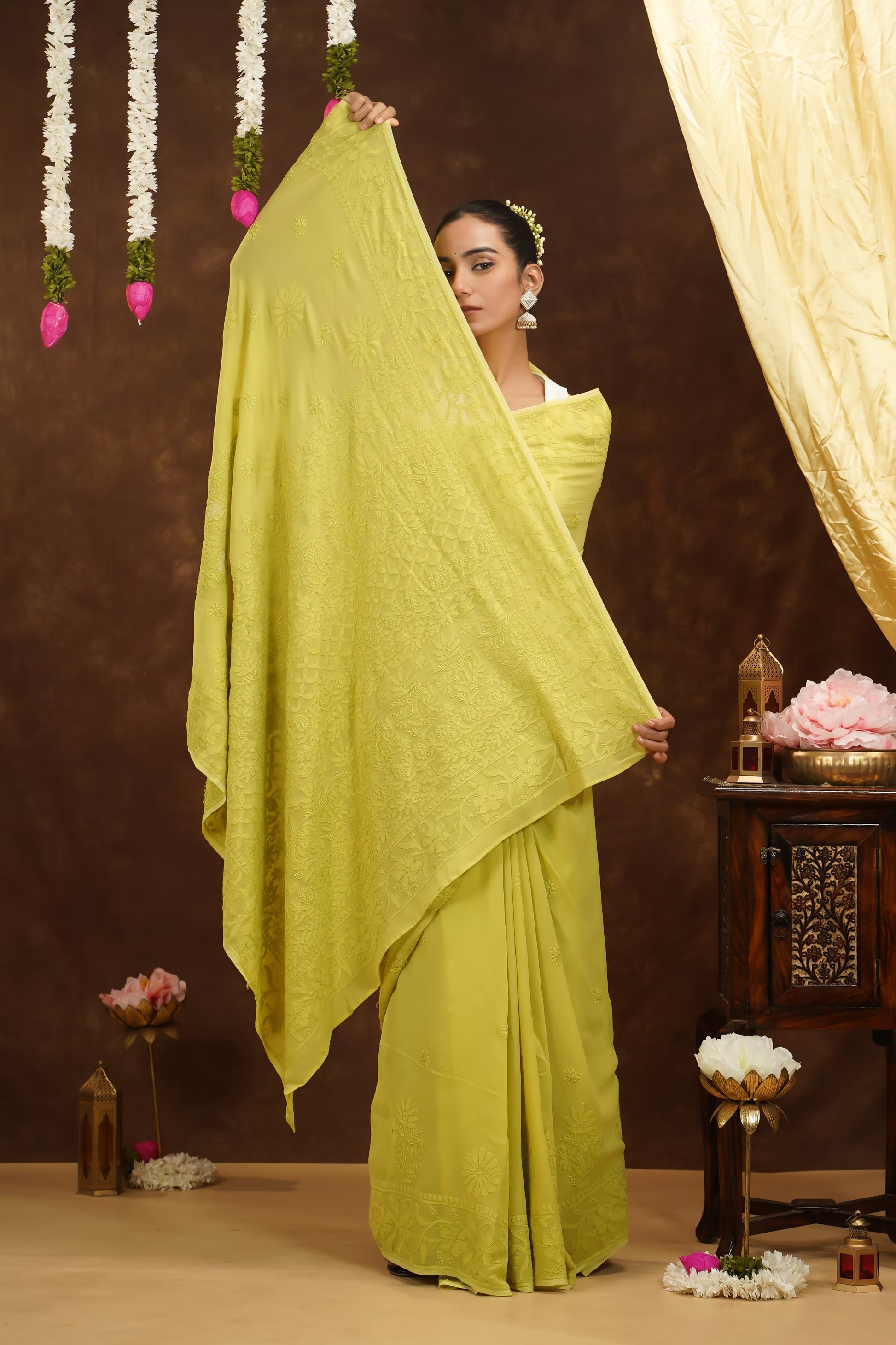 Woman in a bright yellow saree holding it up against a decorated background with flowers and lights.