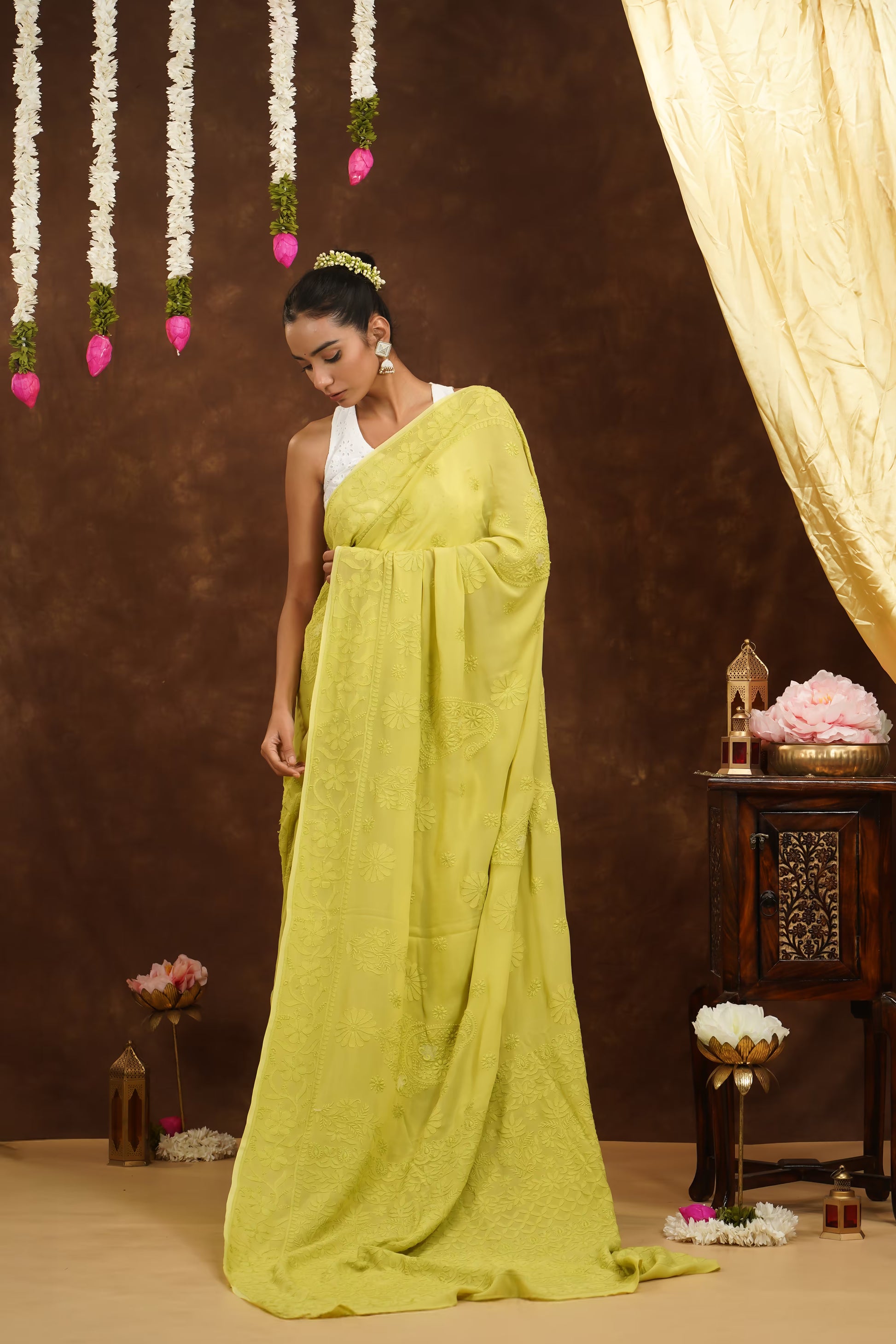 Woman in a bright yellow saree standing in a decorated indoor setting with flowers and lanterns.