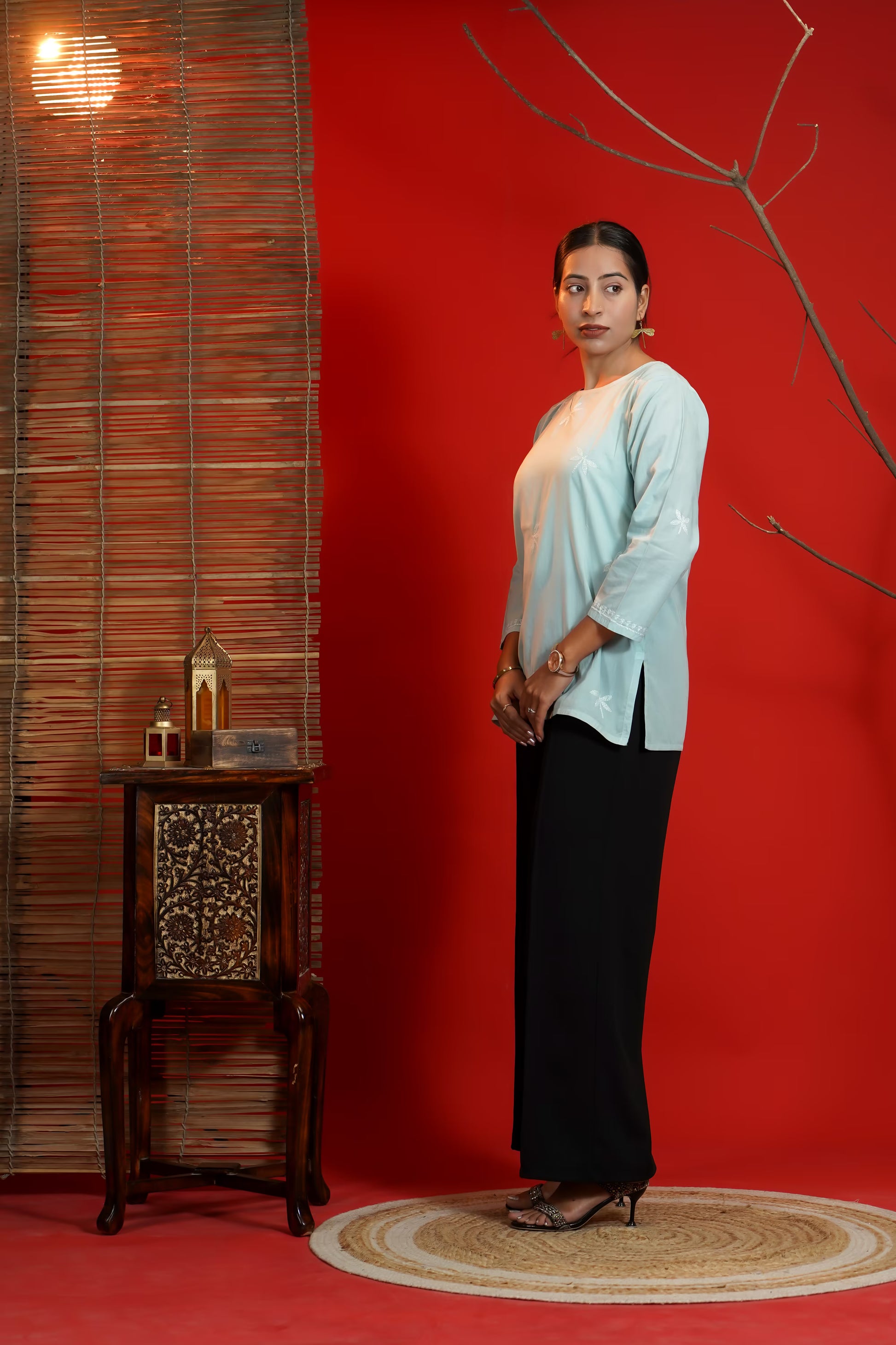 Woman in a Grey top and black pants standing against a red wall with a wooden side table and decorative elements.