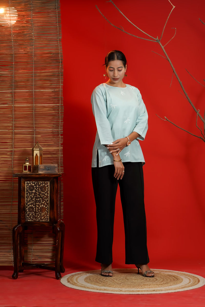 Woman in grey top and black pants standing against a red wall with a wooden side table.