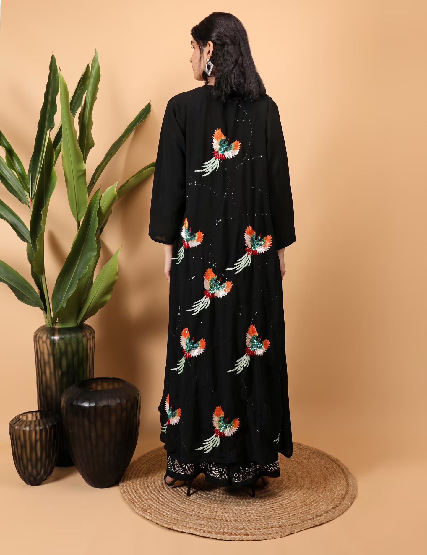 Woman wearing a black Kurti Plazzo Shrug Set with colorful bird embroidery, standing against a beige wall with decorative vases and plants.