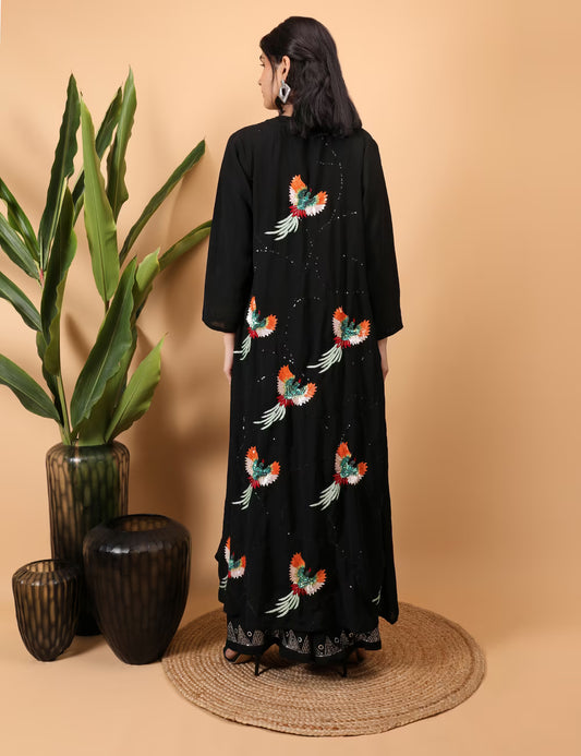 Woman wearing a black Kurti Plazzo Shrug Set with colorful bird embroidery, standing against a beige wall with decorative vases and plants.