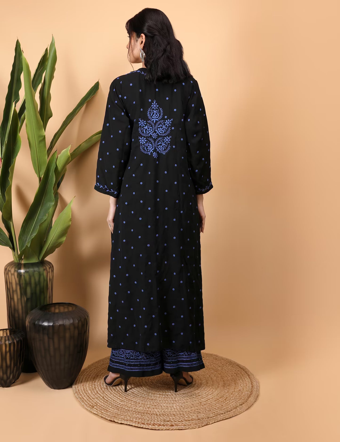Woman wearing a black Kurti Plazzo Shrug Set with blue patterns, standing in front of a beige wall with decorative vases and plants.