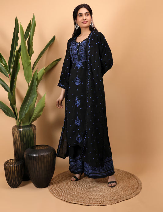 Woman wearing a black and blue embroidered outfit standing next to decorative vases on a beige background
