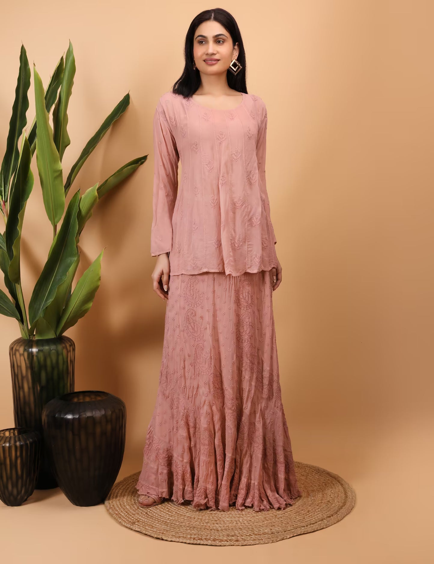 Woman wearing a peach traditional outfit standing against a beige background with decorative vases and plants.