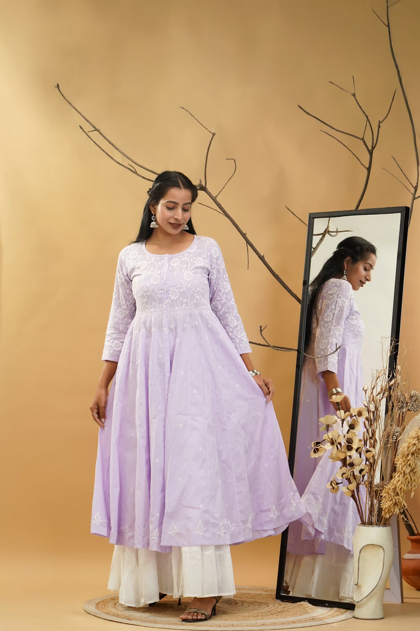 Woman in a light purple dress standing next to a mirror against a beige wall with decorative branches.