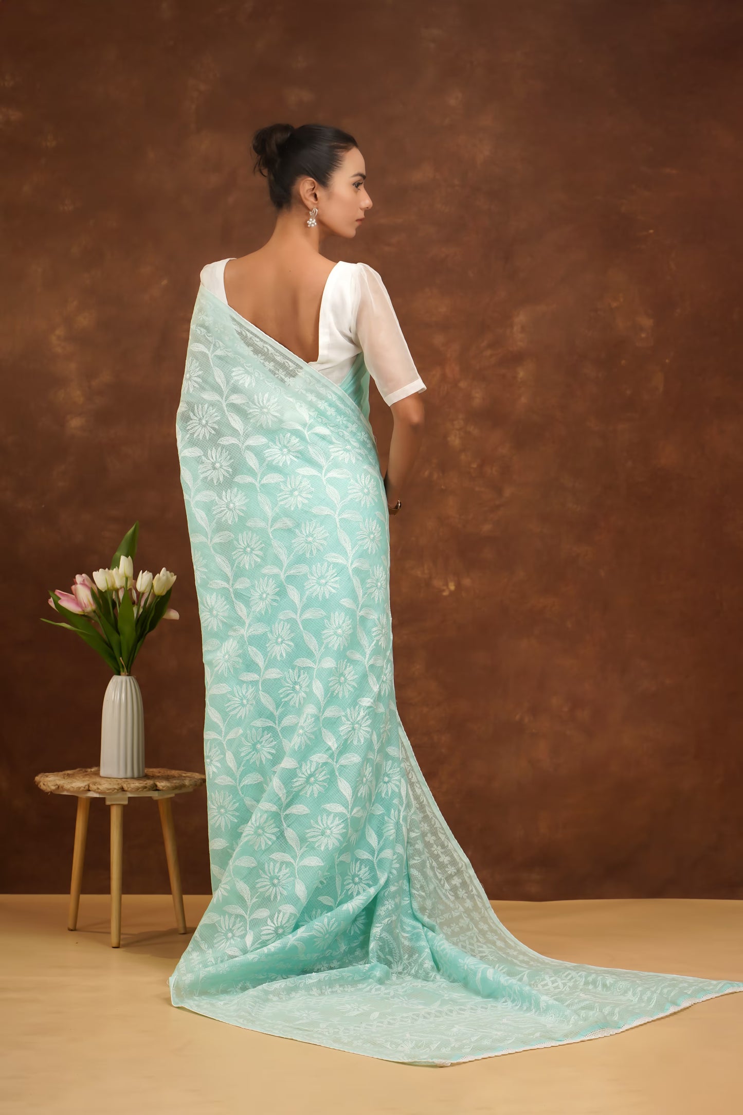 Woman wearing a light blue saree with a white blouse against a brown background