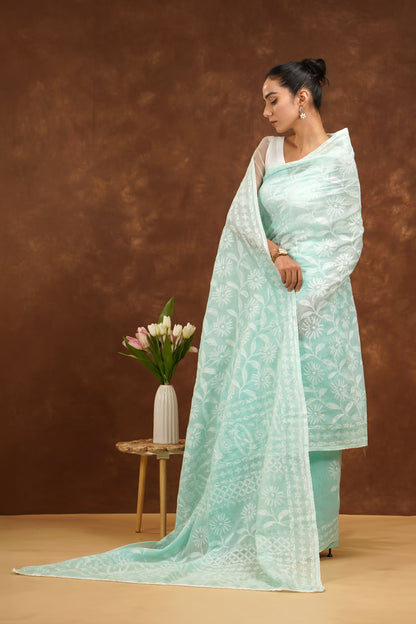 Woman in a light blue saree standing against a brown background with a vase of flowers on a small table.