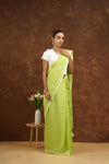Woman in a light green saree standing against a brown background with a small table and flowers.