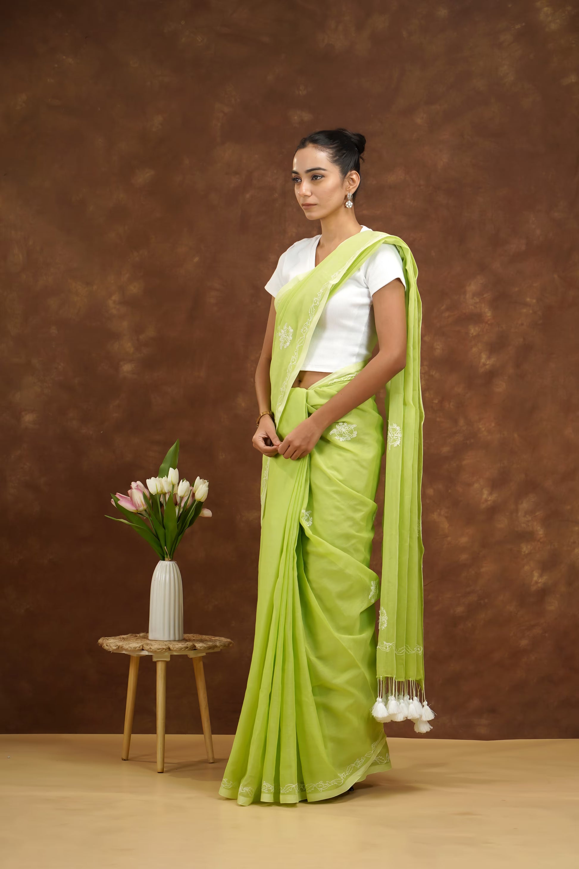 Woman in a green saree standing next to a small table with flowers against a brown background