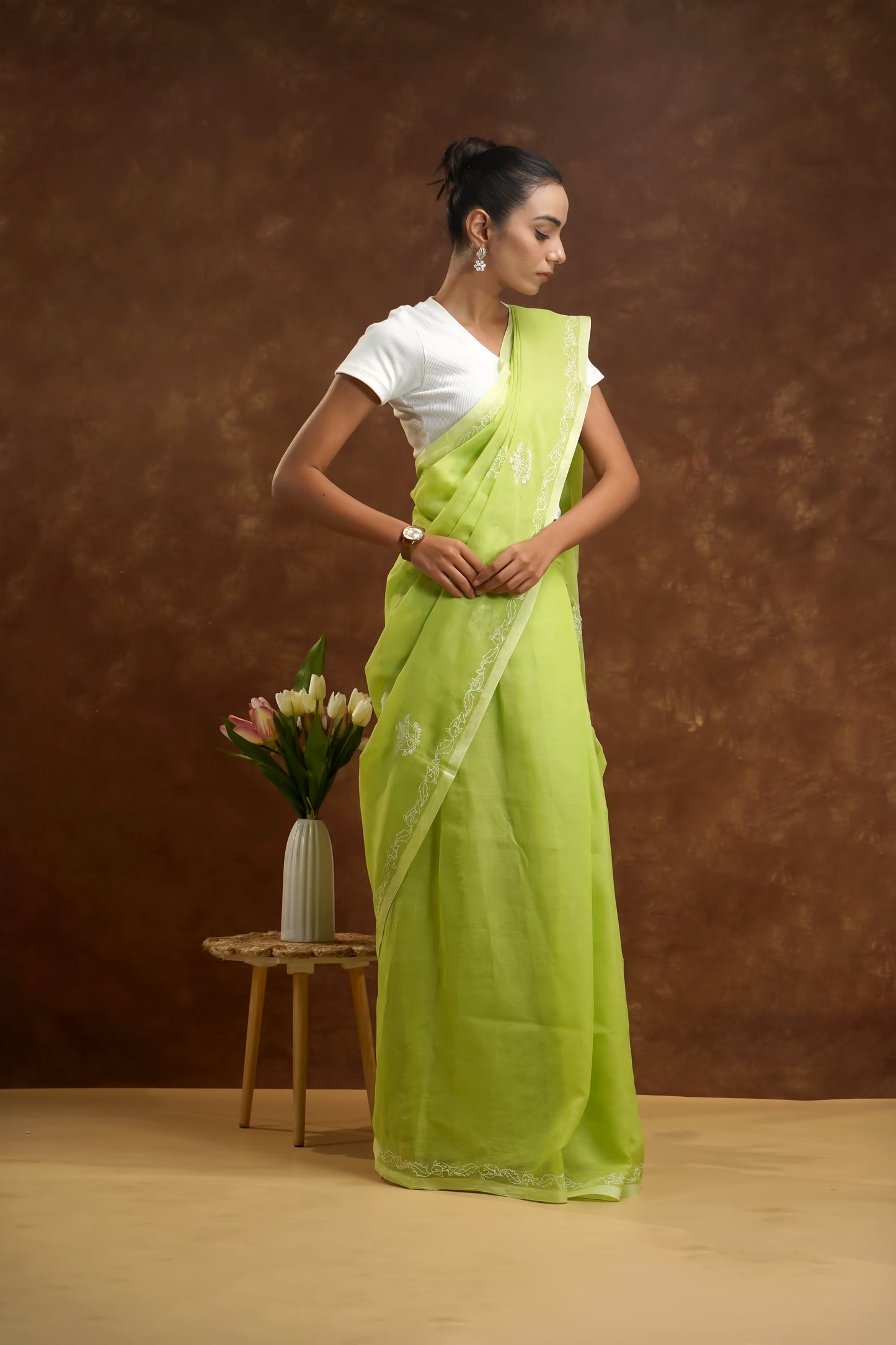Woman in a green saree standing next to a small table with flowers against a brown background