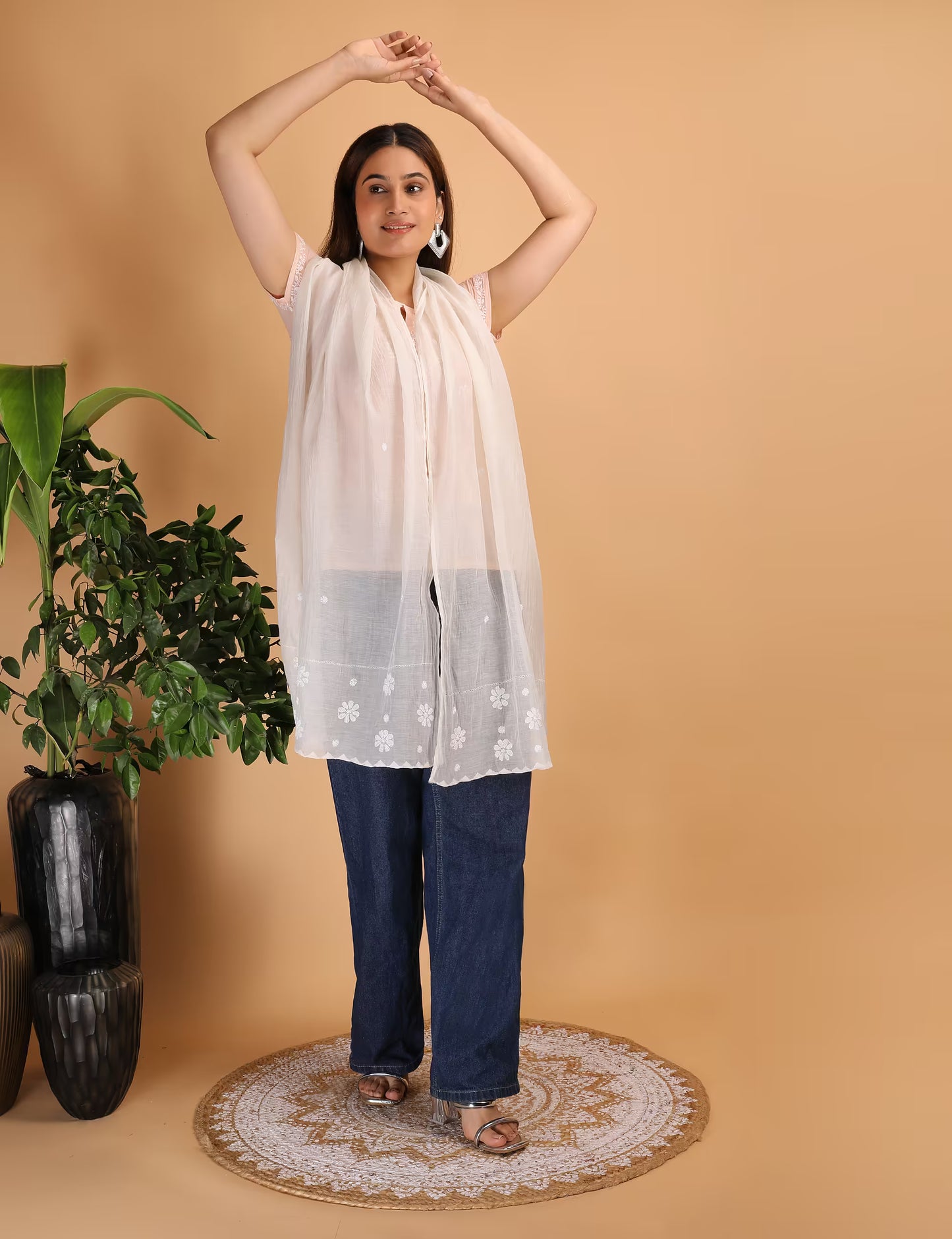 Woman wearing a white Chikankari stole and blue jeans standing against a beige background with a plant to the left.