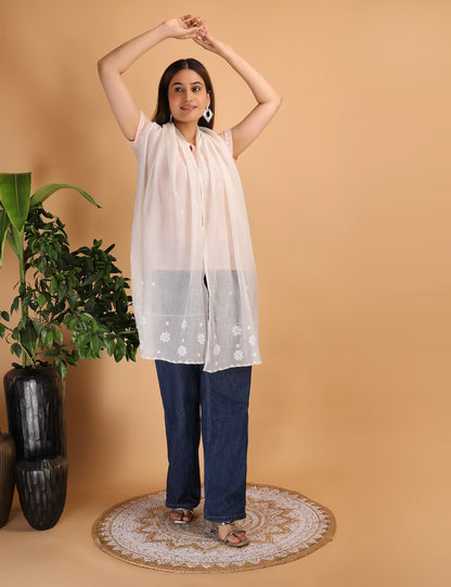 Woman wearing a white Chikankari stole and blue jeans standing against a beige background with a plant to the left.