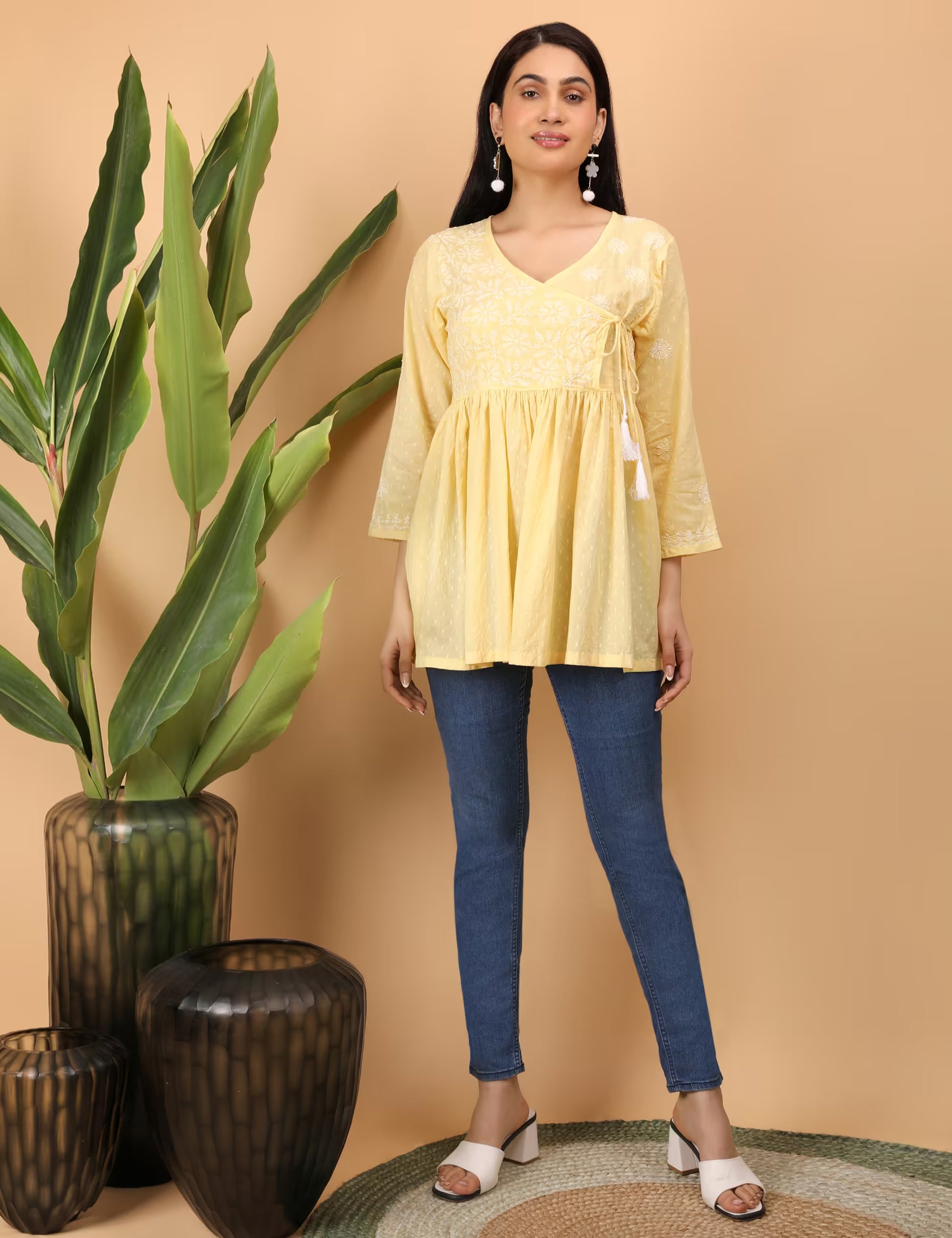 Woman wearing a yellow  Chikankari top and blue jeans standing next to decorative vases with green leaves.