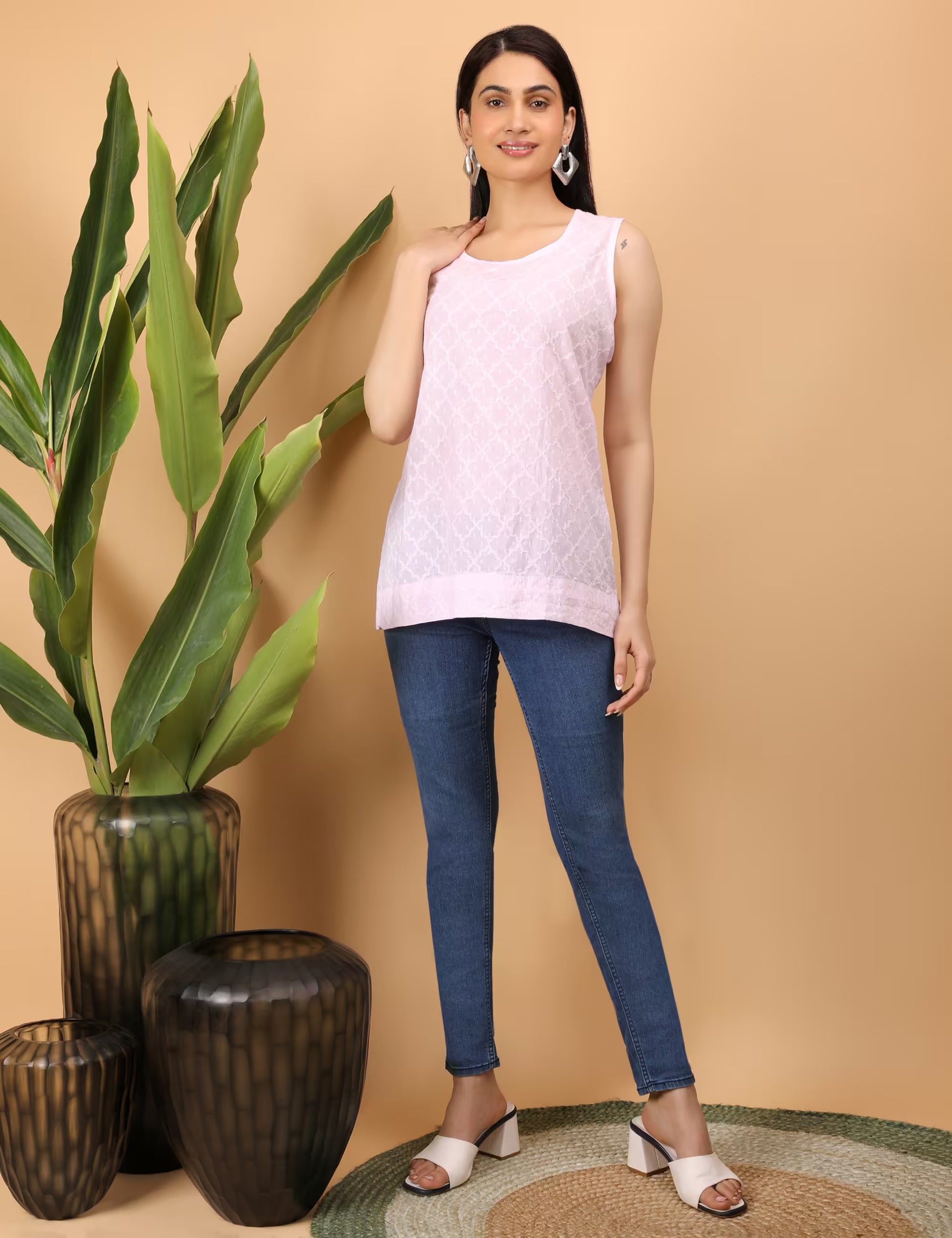 Woman wearing a light pink Chikankari sleeveless top and blue jeans standing next to decorative vases and a plant on a beige background.