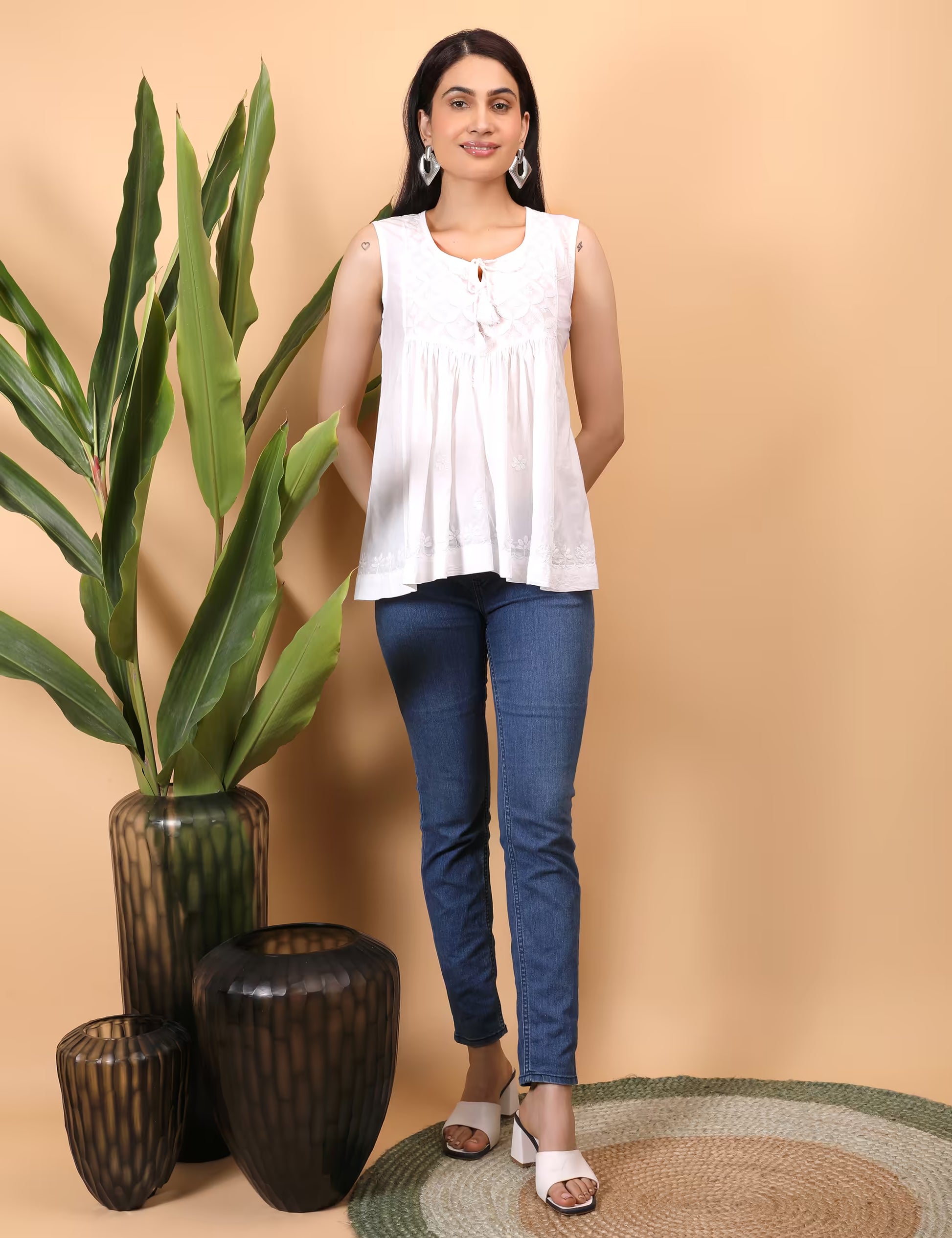 Woman wearing a white sleeveless top and blue jeans standing next to decorative vases with plants on a beige background