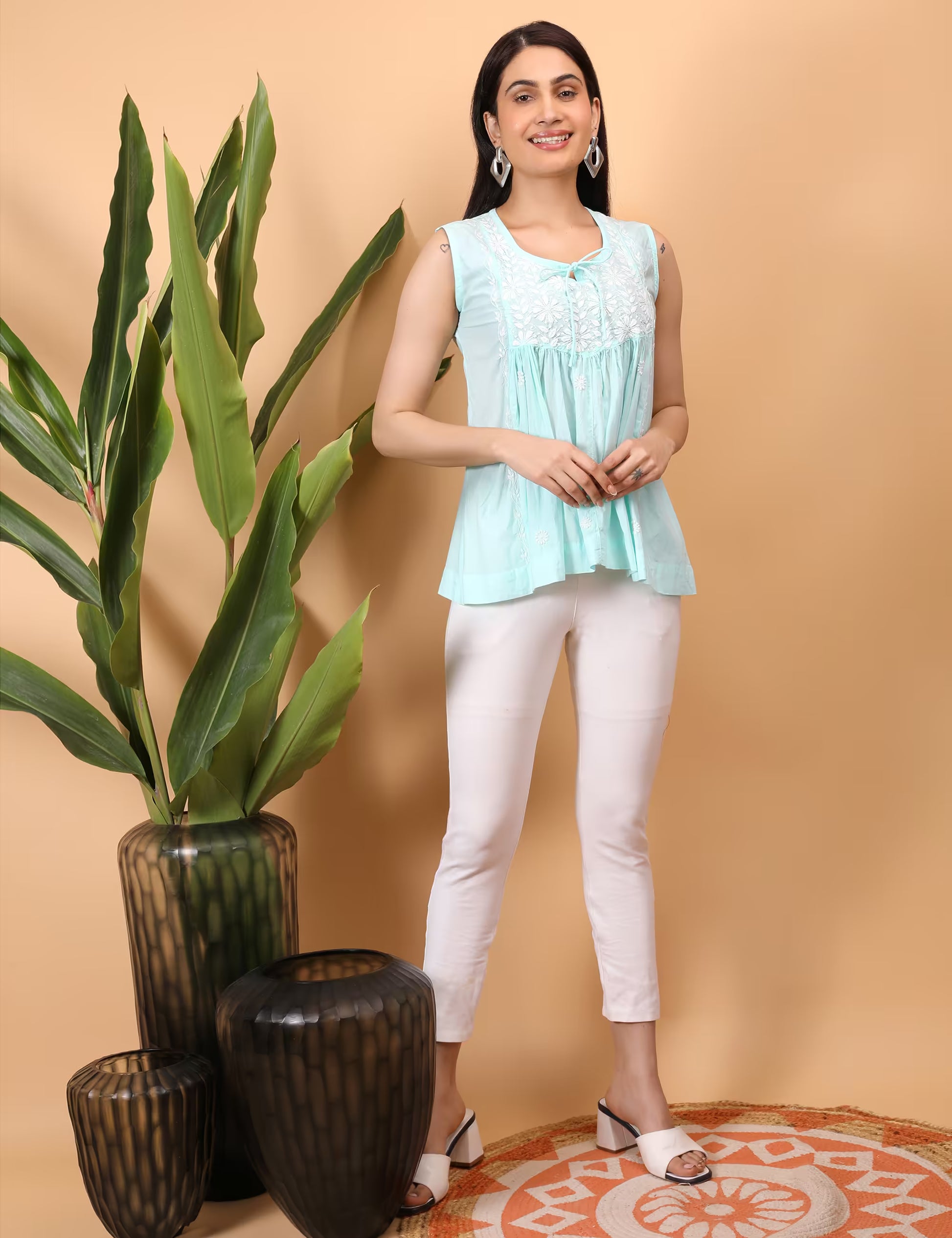 Woman wearing a light blue sleeveless top and white pants standing next to decorative plants on a beige background
