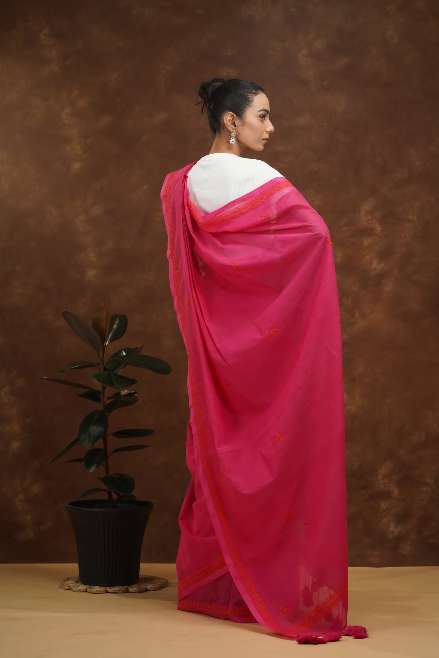 Woman in a megenta saree standing against a brown background with a plant on the left.