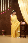 Woman in a yellow saree standing next to a wooden table with decorative items against a brown background.
