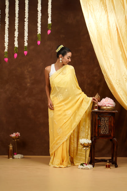 Woman in a yellow saree standing next to a wooden table with decorative items against a brown background.