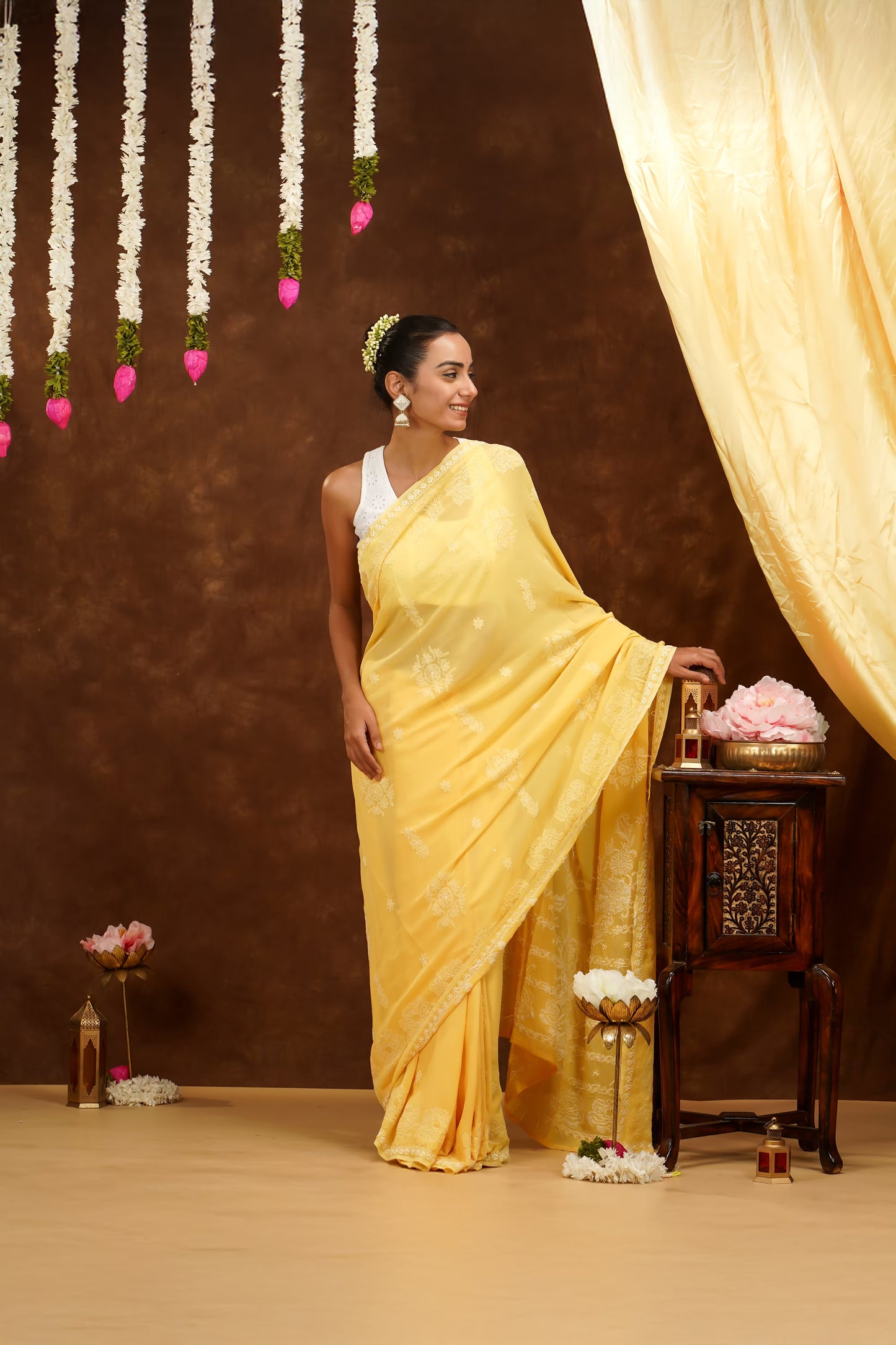 Woman in a yellow saree standing in a decorated room with flowers and curtains.